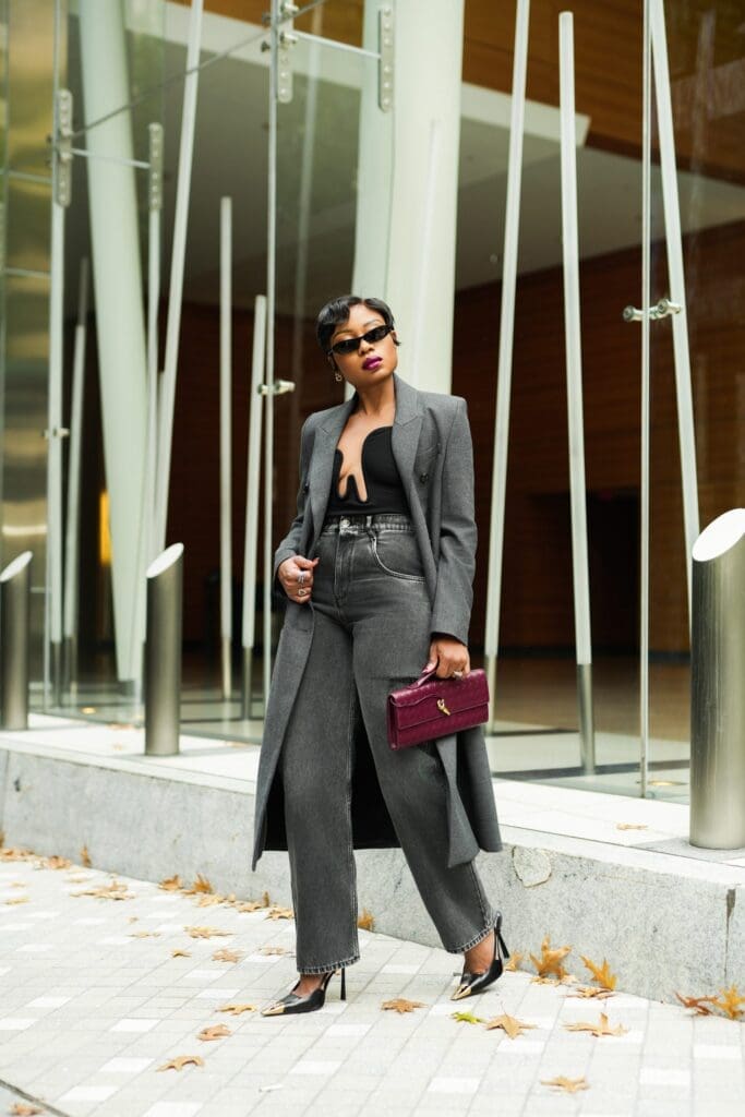 A woman in grey suit and sunglasses holding a purse.