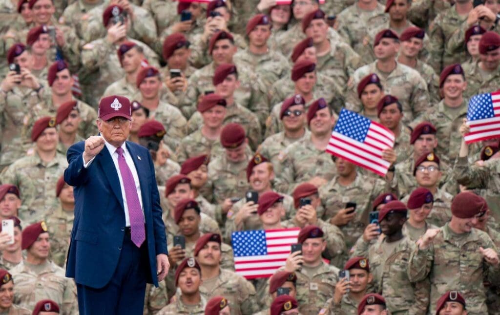 US President Donald Trump wears a baseball cap with military insignia and holds up a fist. He faces away from a crowd of troops at Fort Bragg who wear camouflage-printed fatigues and maroon berets. Some troops hold up their phones or American flags.
