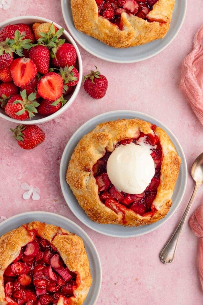 Three mini strawberry rhubarb galettes are on light blue plates, with one topped with a scoop of vanilla ice cream. A bowl of fresh strawberries and a spoon are also visible.
